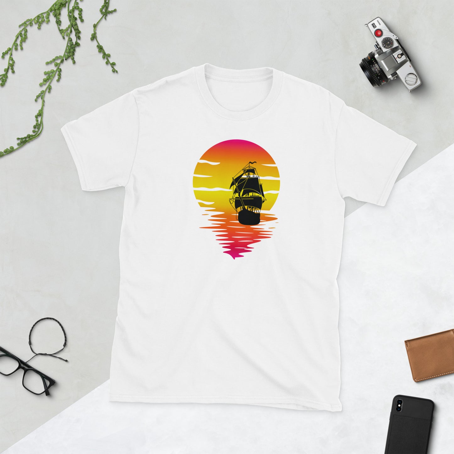 Een unisex basic softstyle t-shirt met een afbeelding van een zonsondergang en een zeilboot op de voorkant, met een bureau waarop camera's en telefoonaccessoires liggen