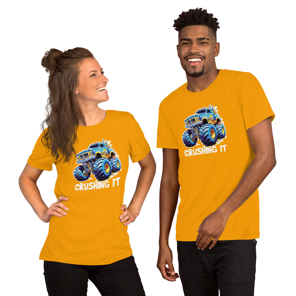 Een goud unisex T-shirt met een afbeelding van een monstertruck, met een lachende man die vooruit kijkt en een lachende vrouw die ook recht vooruit kijkt, tegen een witte achtergrond.