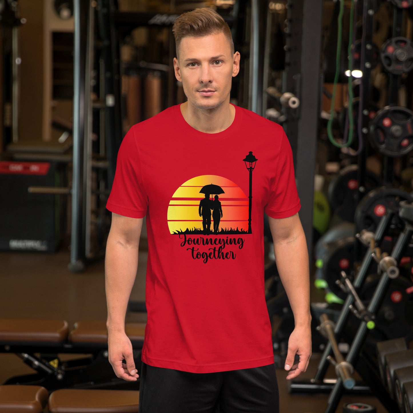 Een unisex rood T-shirt met de titel 'Journeying Together', afbeelding van een koppel hand in hand, gedragen door een man, met een sportschoolachtergrond, vooruitkijkend.