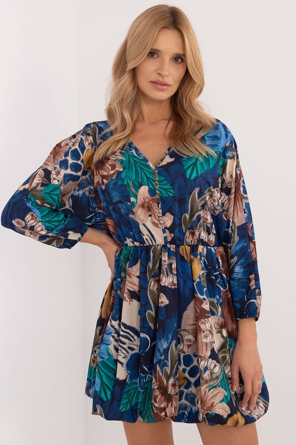 Een vrouw draagt een korte, losvallende jurk met lange mouwen en een opvallende print van kleurrijke bloemen en subtiele dierenmotieven in blauwtinten,voor uitzicht