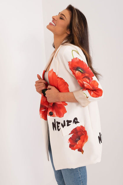 Blije vrouw draagt een witte blazer met rode bloemenprint en zwarte tekst details, gecombineerd met een beige top, met een jeans broek, zij aanzicht
