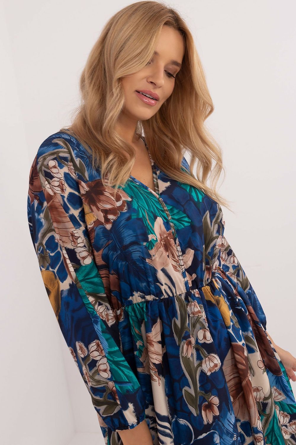 Een vrouw draagt een korte, losvallende jurk met lange mouwen en een opvallende print van kleurrijke bloemen en subtiele dierenmotieven in blauwtinten.links uitzicht