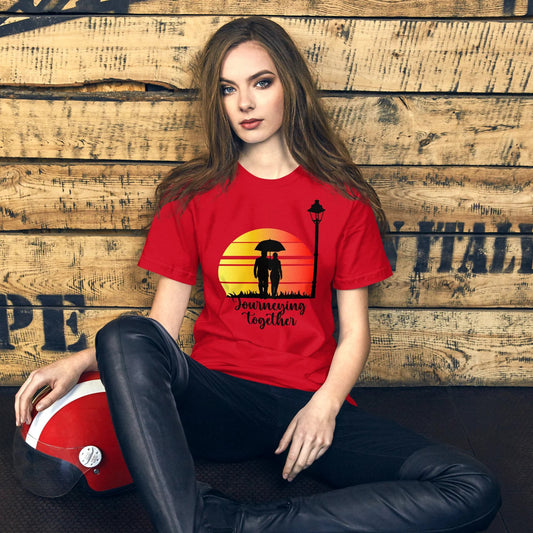 Een unisex rood T-shirt met de titel 'Journeying Together', afgebeeld als koppel hand in hand, gedragen door een vrouw, op een houten achtergrond, vooruitkijkend